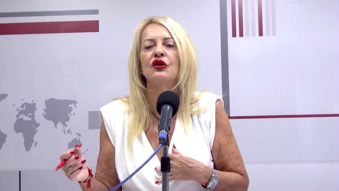 Mujer rubia con camiseta blanca está hablando frente a un micrófono en un estudio con fondo blanco y mapas en gris.