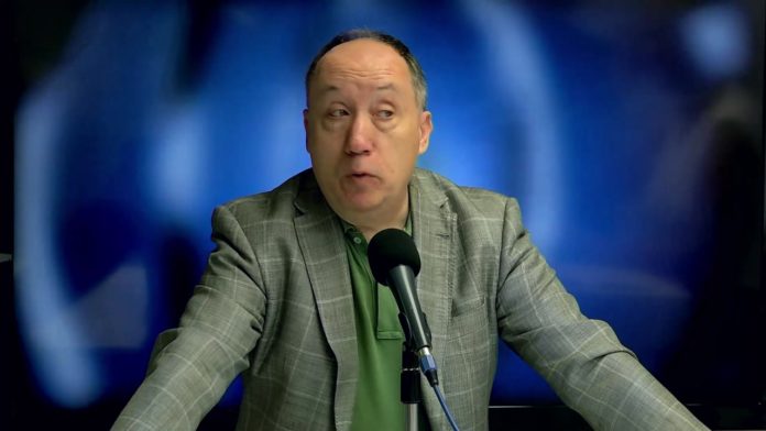 Hombre de mediana edad con chaqueta gris y camisa verde hablando frente a un micrófono negro en un fondo azul desenfocado.