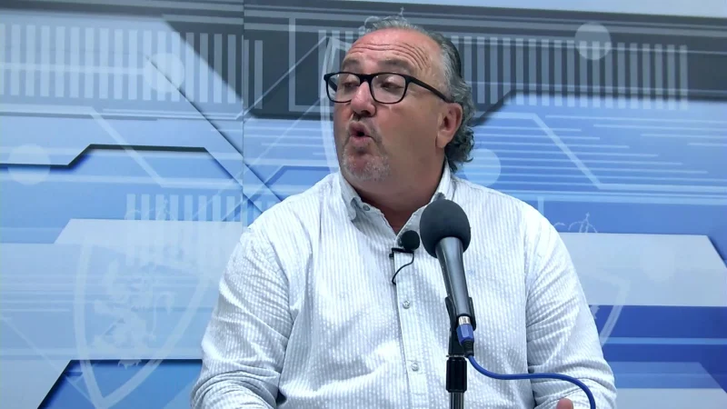 Hombre de mediana edad con gafas y barba canosa, con camisa blanca de rayas, hablando frente a un micrófono en estudio con fondo azul y blanco.