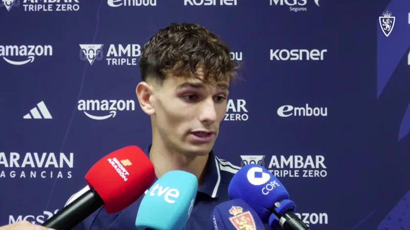 Joven con camiseta azul oscuro habla frente a varios micrófonos de diferentes medios de comunicación en un fondo azul con logotipos.