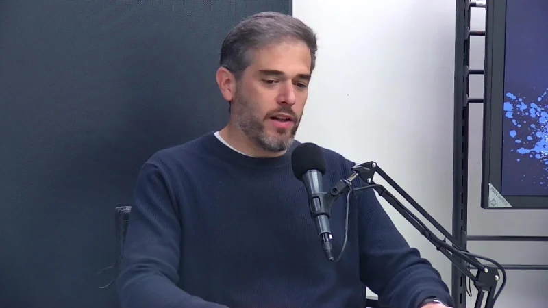 Un hombre con barba y suéter azul está sentado frente a un micrófono de estudio, hablando o explicando algo.