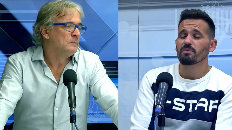 Dos hombres sentados separados por una división, cada uno frente a un micrófono, en un estudio con fondo azul.