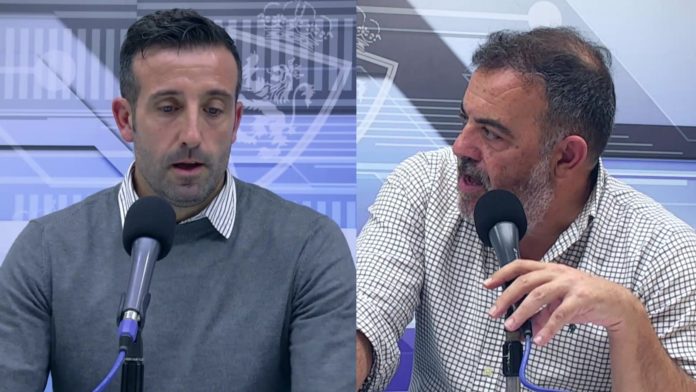 Dos hombres conversan frente a micrófonos en un entorno interior con un fondo azul que tiene un diseño gráfico.