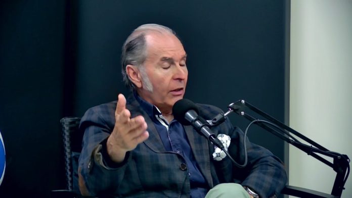 Hombre hablando frente a micrófono en estudio Hombre de cabello canoso y chaqueta a cuadros hablando en un estudio, frente a un micrófono negro montado en un brazo articulado.