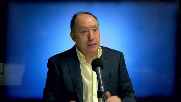 Hombre de mediana edad con chaqueta oscura y camisa clara hablando mientras está sentado frente a un micrófono, con un fondo azul iluminado y difuminado.