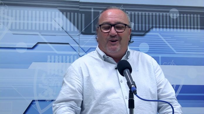 Hombre hablando frente a un micrófono Un hombre de cabello canoso y gafas negras habla frente a un micrófono en un estudio con fondo azul y blanco con figuras geométricas.