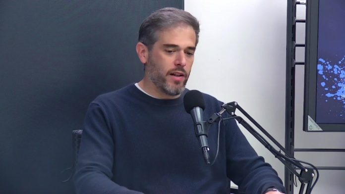 Un hombre con barba y suéter azul está sentado frente a un micrófono de estudio, hablando o explicando algo.